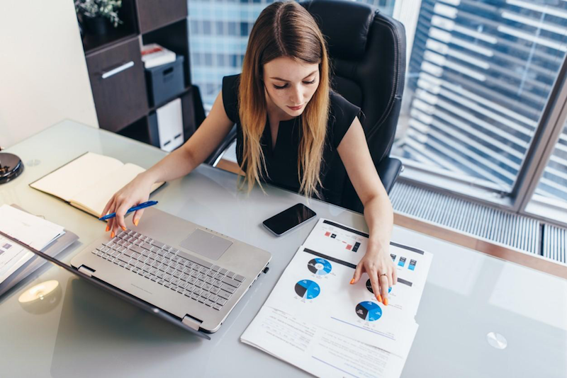 A female business analyst sits at desk in front of laptop looking at printed pie charts showing data.