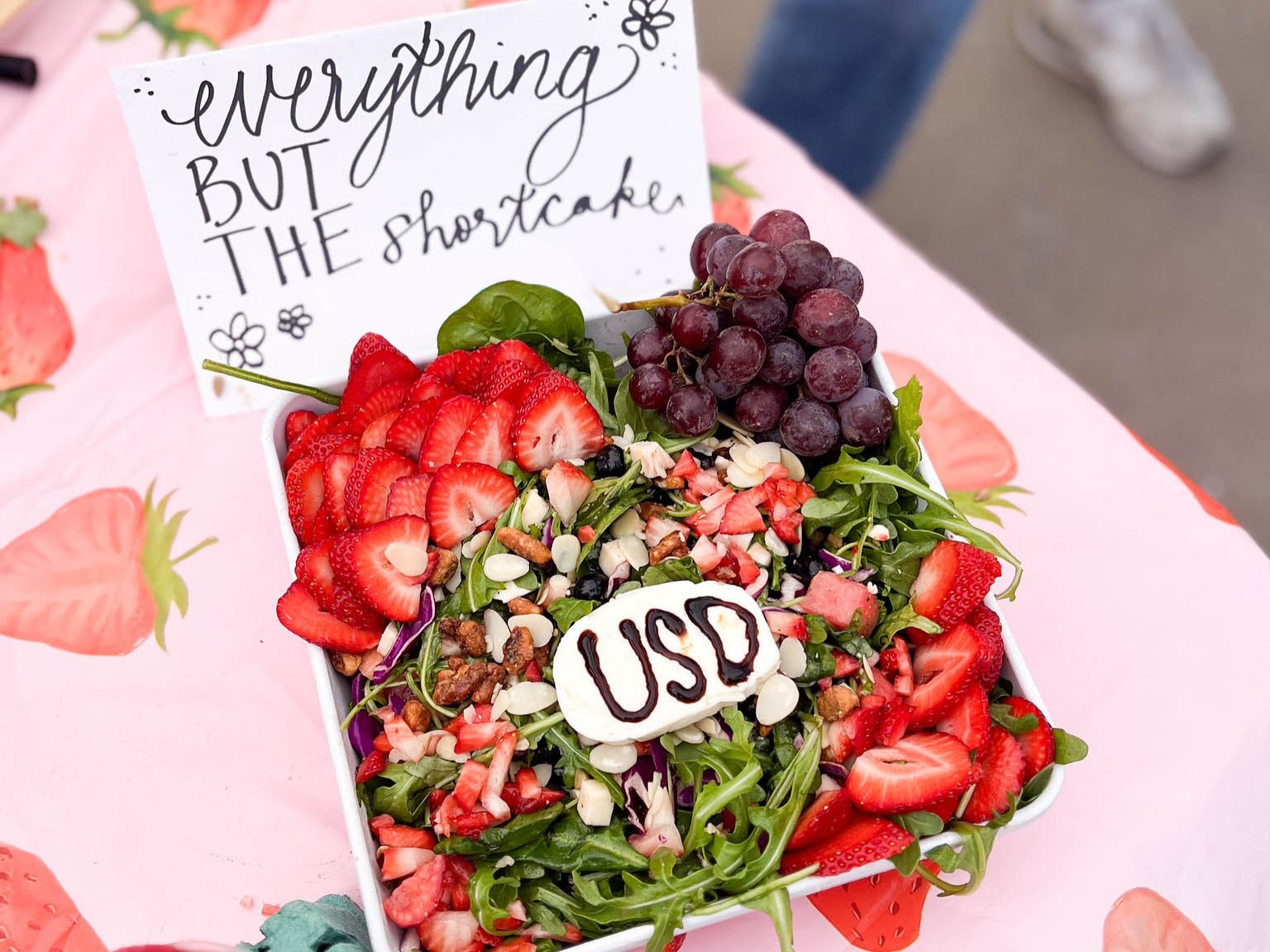 A beautiful salad with greens and strawberries and USD written in balsamic vinegar on a slice of mozzarella cheese. And a sign that says "Everything but the shortcake."