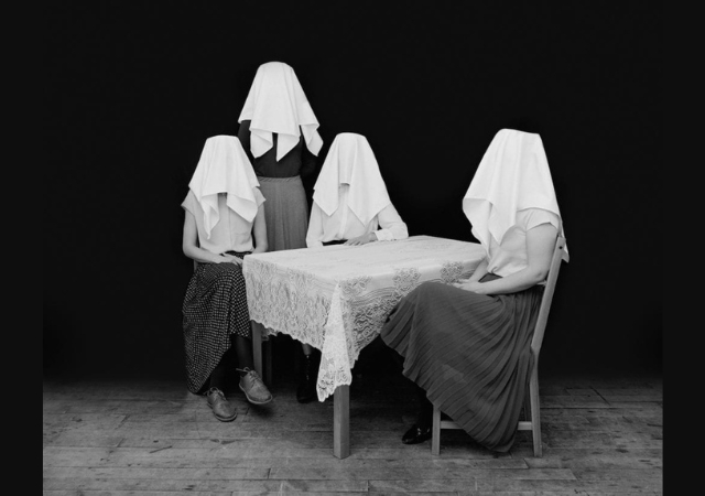 This is a black and white image of four women sitting at a table with white cloths over their heads.