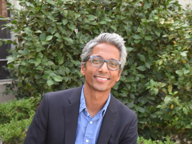 Dr. Devadoss is smiling, sitting in front of a green …