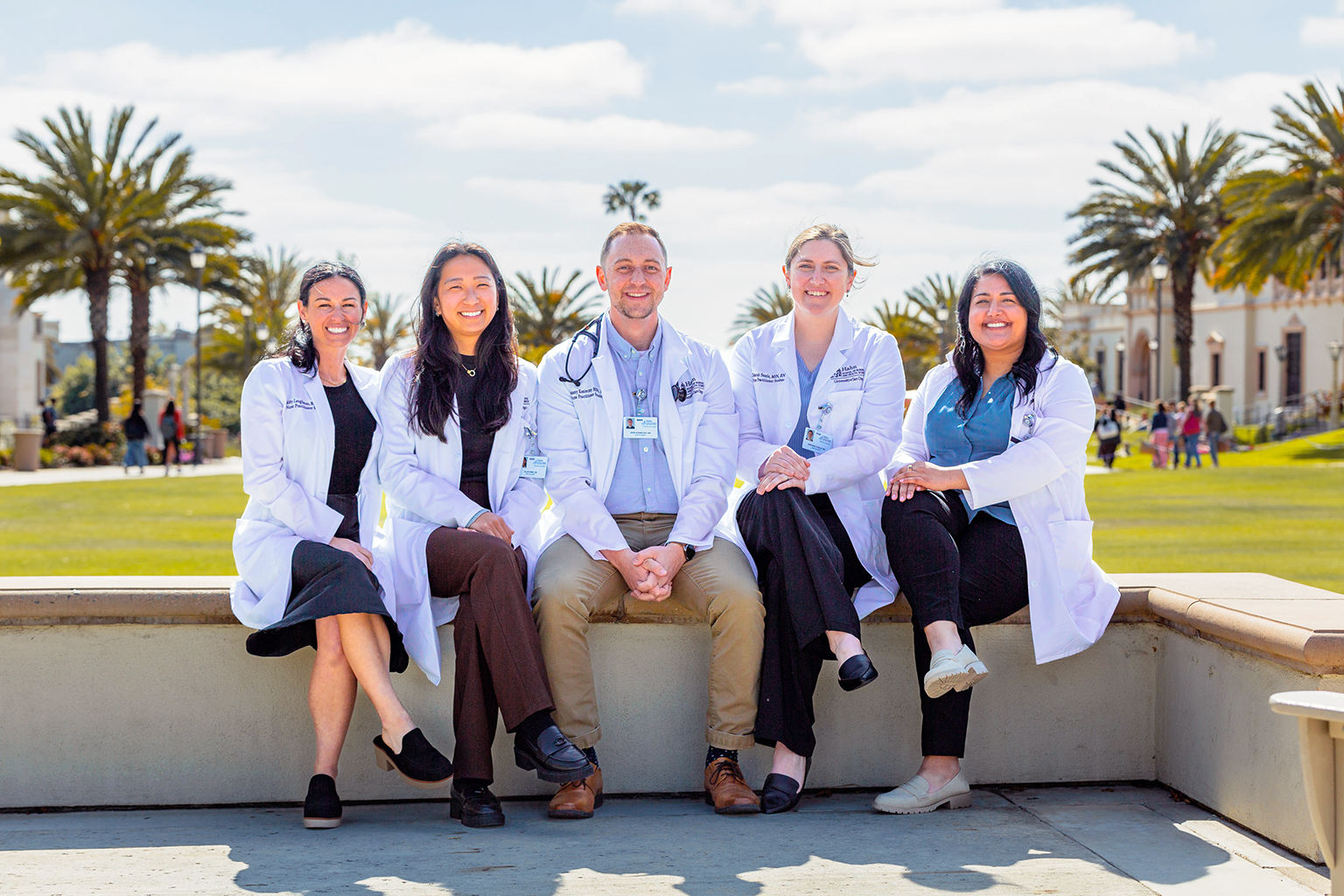 Five University of San Diego Hahn School of Nursing nurse practitioner students wearing white lab coats sit together on campus with palm trees and campus buildings in the background.