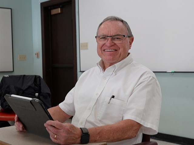 Roger is sitting at a desk in a classroom, smiling and holding onto a computer tablet. A white board is behind him.