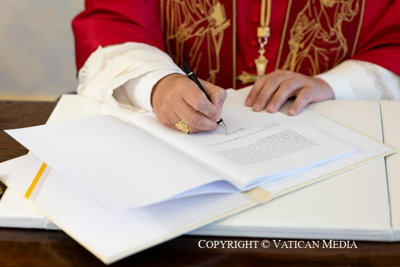 This is a close up photograph of Poe Leo XIV signing the encyclical. The image focuses on his hands, holding a pen and leaning on a desk with the letter.