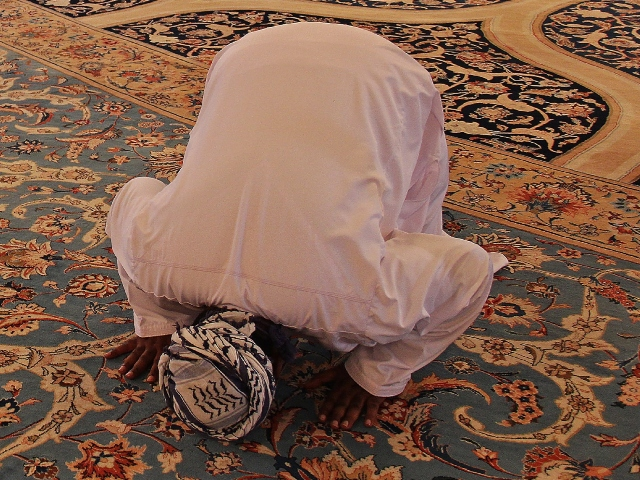 Photo of a man in prayer position. He is wearing a white tunic and headrag. He is bending over at the hips, with both hands and his face toward an elaborately decorated rug.