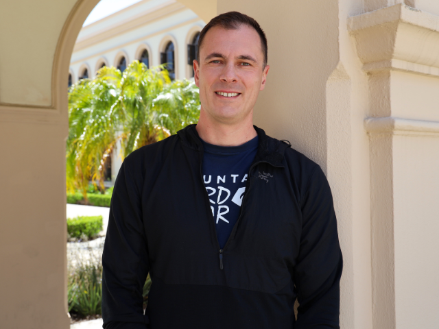 Erik Christensen stands in between an archway in the courtyard between Camino and Founders. A palm tree peaks out from behind his left shoulder.