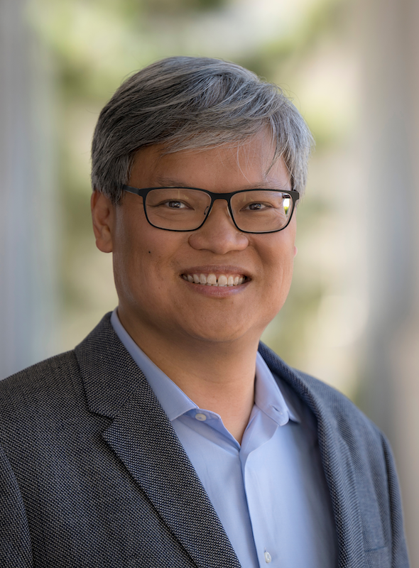 A person wearing a textured gray blazer over a light blue button-up shirt, standing in a softly lit outdoor setting with a neutral background featuring soft greenery tones. The attire and setting convey a professional, academic context suitable for a faculty profile photo.