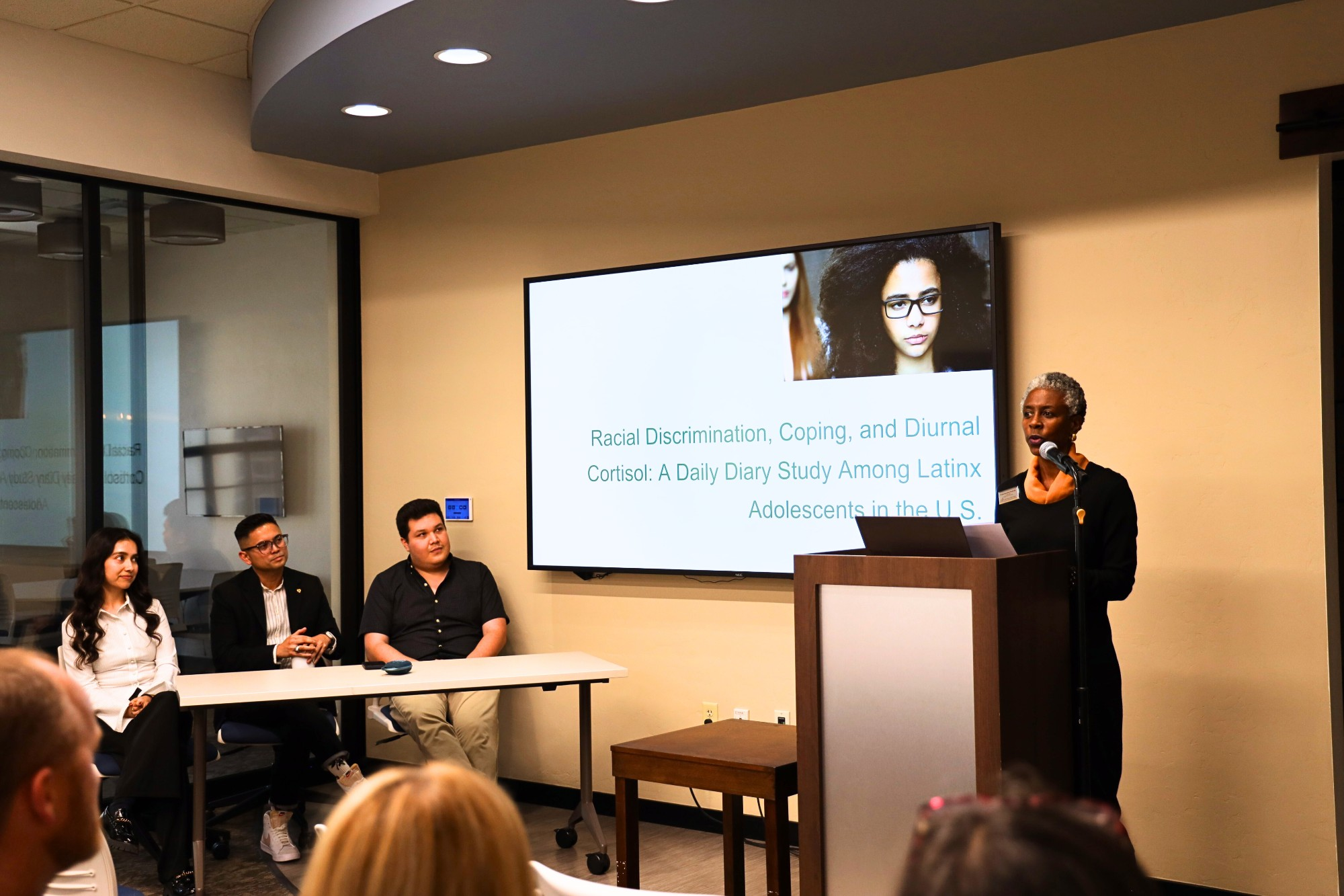 Pauline Berryman Powell standing at a podium introducing the event while Dr. Sarsar, Paloma and Dr. León Sandoval look on.