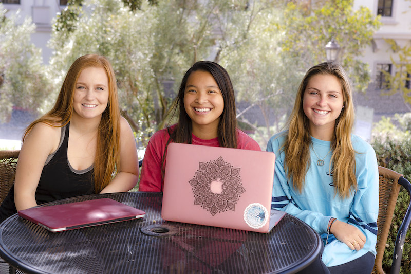 Three undergrad females sit smiling in front of a laptop outdoors at the University of San Diego