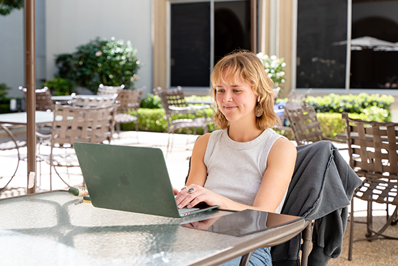 Nursing student sitting outdoors at a patio table, smiling while using a laptop on the University of San Diego campus.