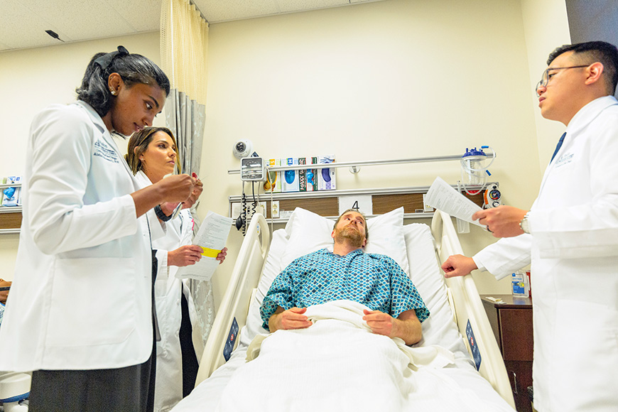 Three nursing students in white clinical coats stand around a patient bed in a high-fidelity simulation lab, reviewing charts and preparing for a medical procedure.