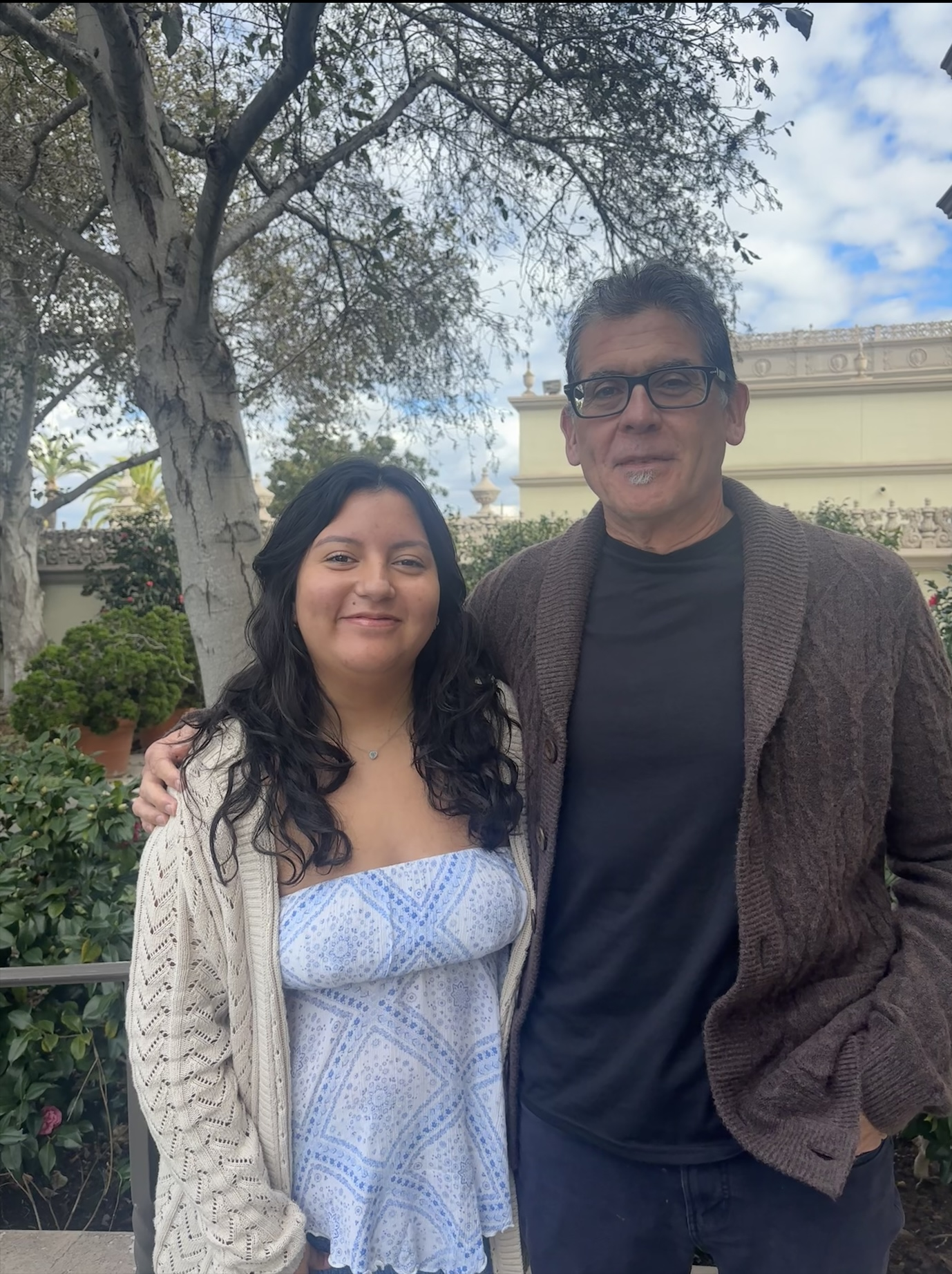 Jennifer Torres Leon and Dr. Cid Martinez are standing outdoors in front of trees and a light-colored building. They are smiling at the camera, and one has their arm around the other. The setting appears calm and pleasant, with greenery and a partly cloudy sky in the background.