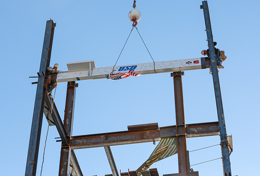 Two construction workers work together to secure a historic beam into place atop the frame of USD's  new Wellness Center and Basketball Practice Facility during a topping off ceremony on Monday, Jan. 29, 2024. (Photo by Matthew Piechalak)